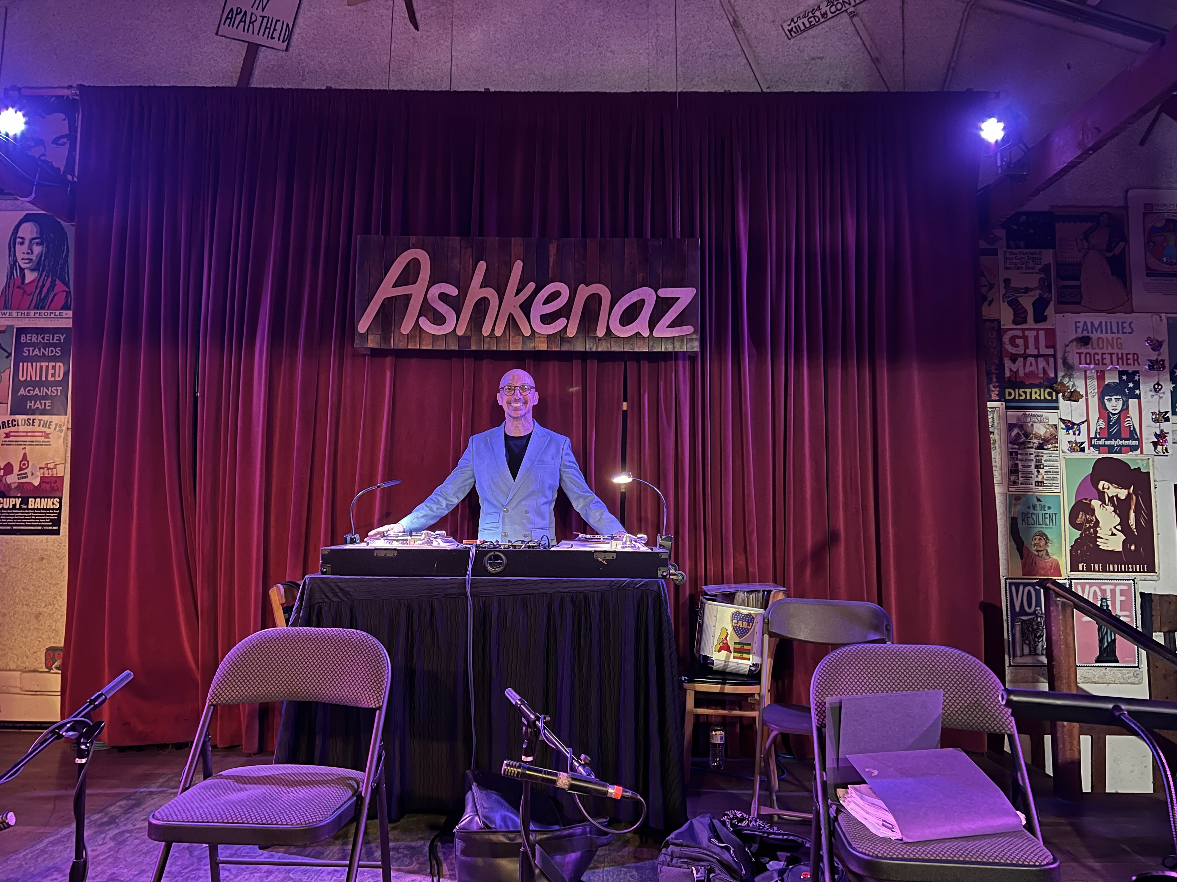 DJ Charles Carbone standing at the turntables on the Ashkenaz stage beneath the venue sign.